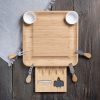 A bamboo cheese board with utensils and two small bowls set on a wooden table with a gray napkin.