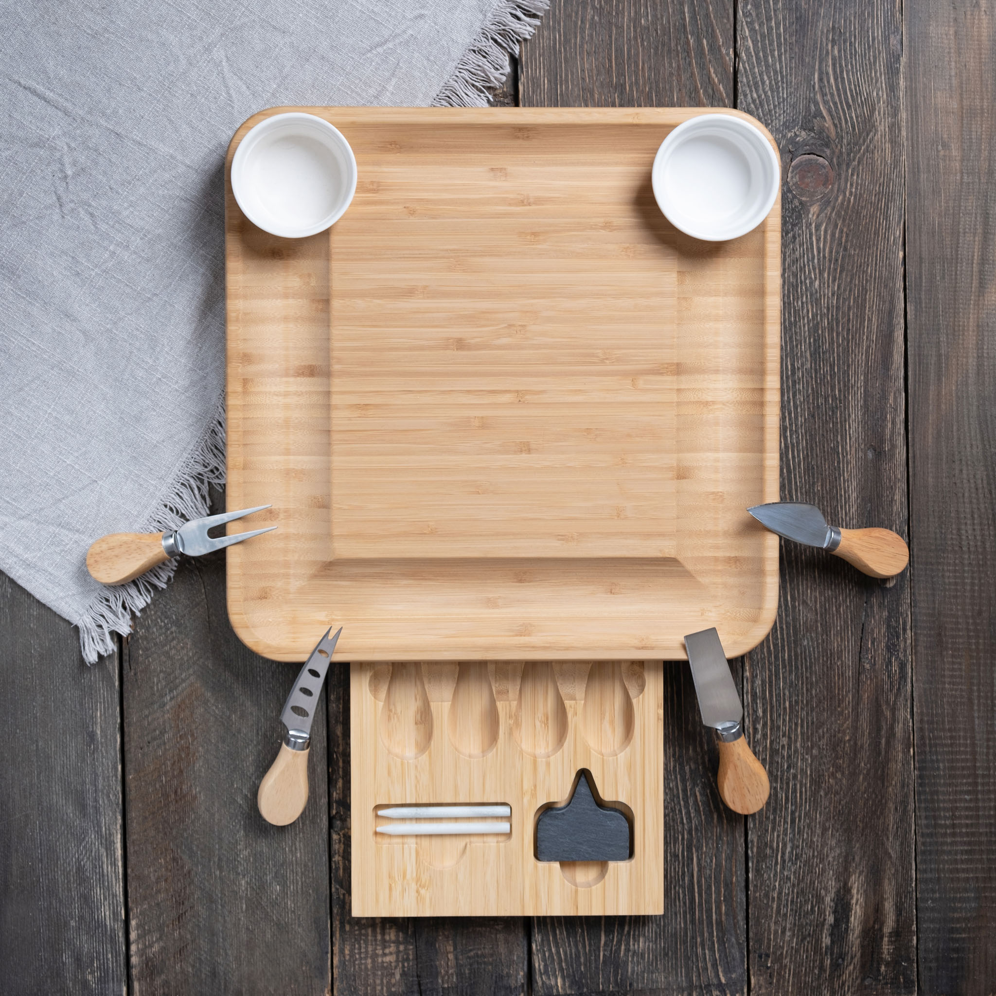 A bamboo cheese board with utensils and two small bowls set on a wooden table with a gray napkin.