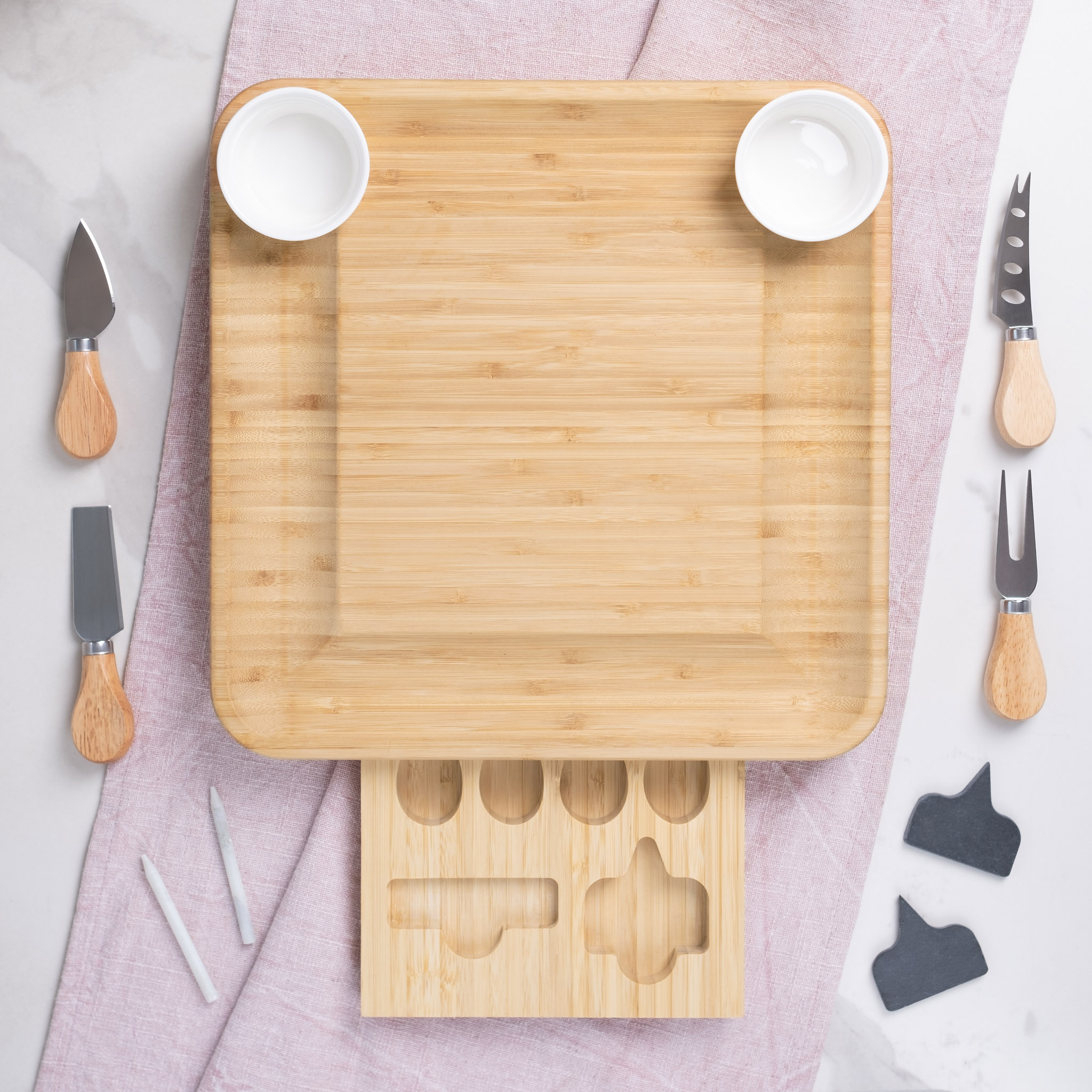 A neatly arranged cheese board with an assortment of knives and bowls on a marble background.