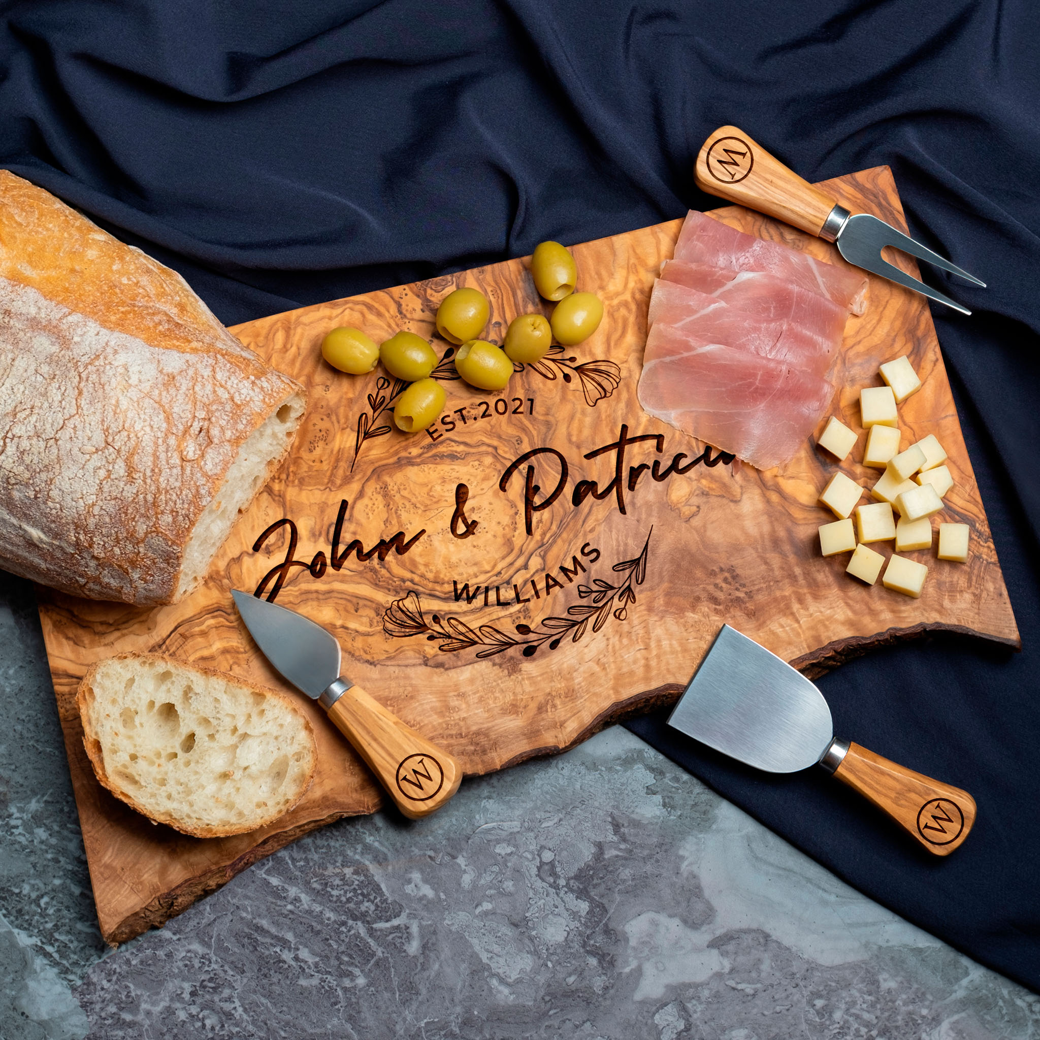A personalized wooden cheese board with utensils, served with bread, olives, prosciutto, and cheese cubes.