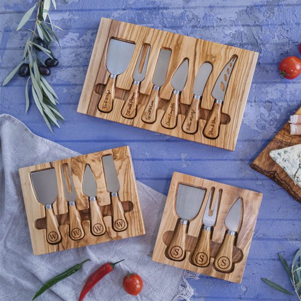 A wooden cheese board with cheese knives and olives.
