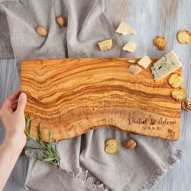 A hand holding a personalized wooden cheese board with assorted cheese pieces, crackers, and a sprig of rosemary on a gray fabric surface.