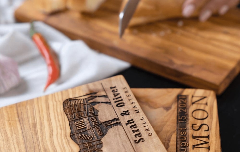A person slicing vegetables on a wooden cutting board with a personalized engraving, "sarah & oliver, august 15, 2021.
